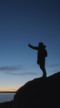 Silhouette of a man standing on a rocky shore and taking a selfie with a smartphone at twilight. Dark figure against a deep blue evening sky and calm water creates a dramatic outdoor travel scene with