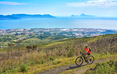 Fototapeta premium Smiling senior woman riding an electric mountain bike above Pompeii. Panoramic view of towns, mountains, and the Gulf of Naples, Active lifestyle, and healthy aging in Pompei, Campania, Italy. 