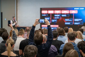 Audience captures presentation on smartphone during busy business conference, workshop slide visible on large screen. © Anton Gvozdikov