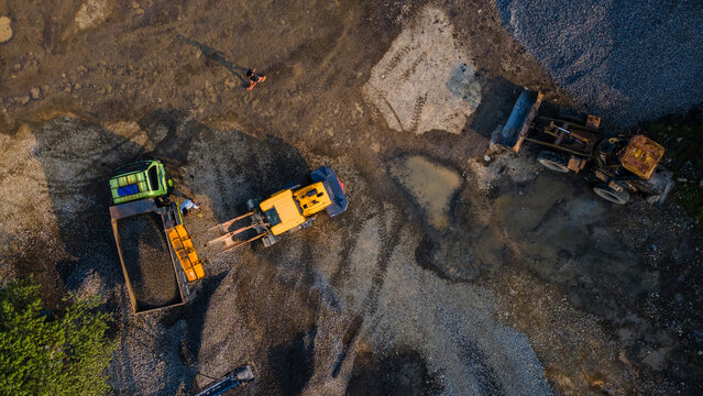 Aerial view of heavy machinery including an excavator and a truck working on a gravel construction site with workers and puddles in Karanglewas, Central Java, Indonesia.