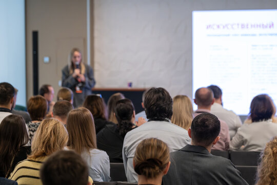 Audience listening to business presentation at professional conference, speaker using projected slides, focused attendees taking notes and engaging with topic.