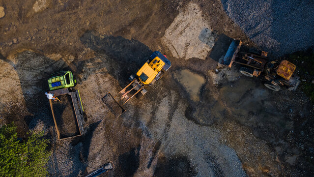 Aerial view of heavy machinery including an excavator and trucks working at a construction site with piles of gravel and muddy terrain in Karanglewas, Central Java, Indonesia.