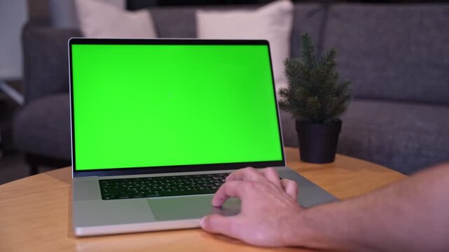 Close-up shot of a person's hand using the trackpad of a laptop with a blank green screen. The computer rests on a wooden table in a softly lit, modern living room with a blurred sofa background.