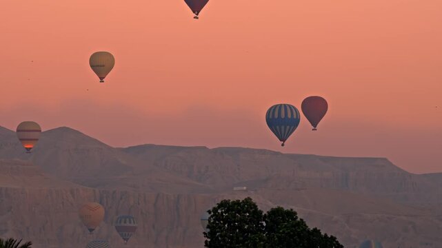 A wide, sweeping view capturing a fleet of hot air balloons floating over the dramatic mountain ridges of the Valley of the Kings in Luxor during a stunning golden sunset.