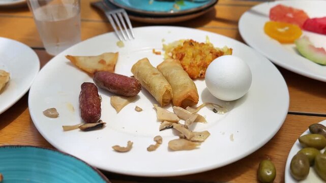 Close-up shot of a partially eaten breakfast plate on a wooden table in bright daylight. The white plate contains a boiled egg, sausages, spring rolls, and hash browns, surrounded by fruit and olives.