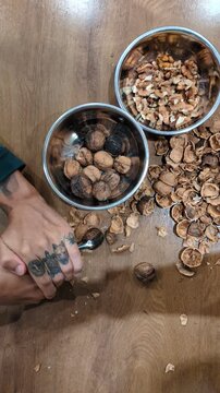 Top-view vertical shot of a person with tattooed hands using a metal nutcracker tool to crack fresh organic walnuts on a rustic wooden table surrounded by healthy kernels and broken shells.