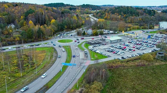 Aerial drone view of a busy highway intersection and roundabout on the E6 motorway in Sweden, featuring a restaurant and service area during autumn with colorful forest landscape.