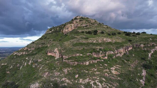 Aerial drone footage flying backward from Cerro del Picacho, revealing the surrounding landscape beneath dramatic storm clouds in Aguascalientes, Mexico. Cinematic mountain scenery with expansive view