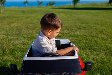 Fototapeta premium Boy sitting in red electric ride on toy car on green lawn looking sideways with blue sea on horizon