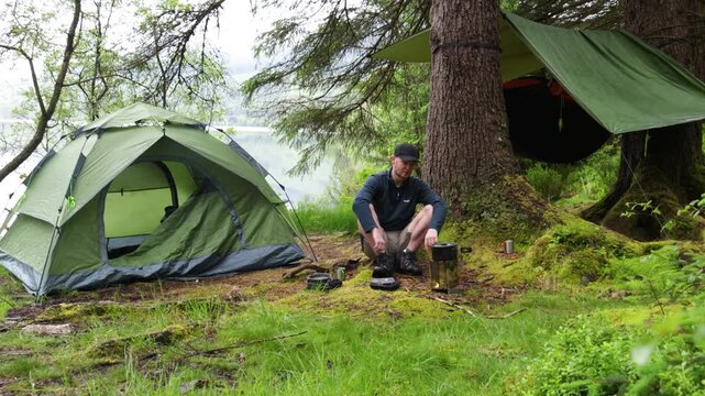 Camper waiting for meal to cook on small campfire stove beside tent by a quiet lake, remote forest campsite with tarp shelter and backpacking gear.