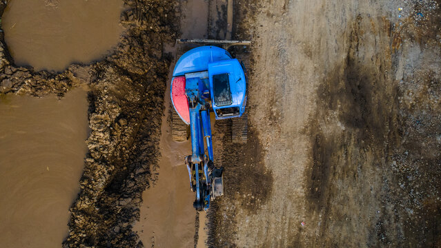 Aerial view of a blue excavator operating on muddy ground at a construction site with industrial machinery and earthmoving equipment in Karanglewas, Central Java, Indonesia.