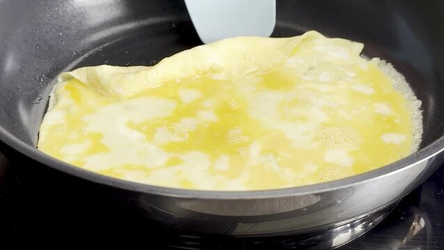 Cooking process of a golden omelette in a frying pan, spatula lifting edge while egg mixture bubbles and cooks, showcasing gradual folding technique in a kitchen setting