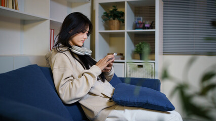 Fototapeta premium Young woman sitting on sofa at home using smartphone with headphones smiling near indoor plants and pillows in living room, lifestyle and technology concepts