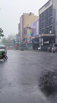 Jakarta, Indonesia - Heavy rain pouring on a busy city intersection with motorcycles, a blue bajaj, and vehicles driving through the wet street.