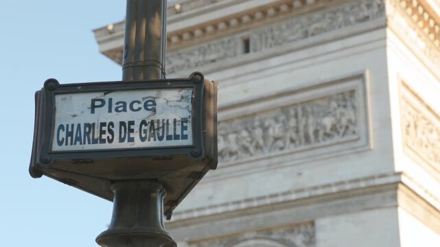 Close-up of a classic Parisian street sign reading Place Charles de Gaulle on a lamppost, with the detailed carvings of the historic Arc de Triomphe monument blurred in the background under a clear bl