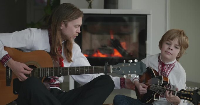 Man and Kid Boy Play Music on Guitar and Mandolin Sitting on Floor Near Fireplace. 4K 10-bit