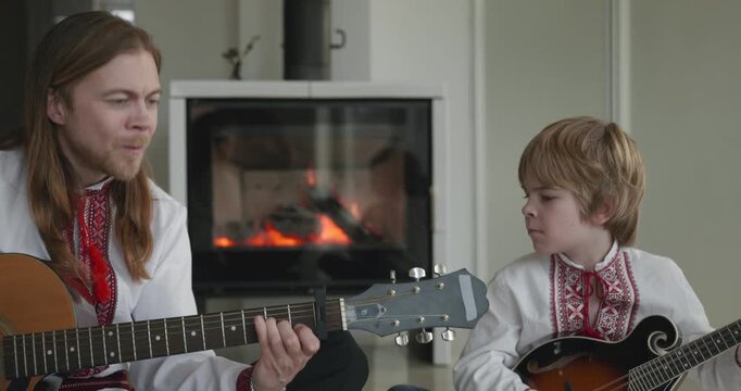 Man and Kid Boy Play Music on Guitar and Mandolin Sitting on Floor Near Fireplace. 4K 10-bit