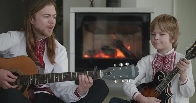 Man and Kid Boy Play Music on Guitar and Mandolin Sitting on Floor Near Fireplace. 4K 10-bit