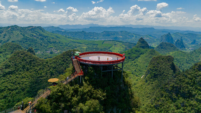 Aerial view of the circular red observation deck at Ruyi Peak ropeway scenic area overlooking lush karst mountains and green hills under a blue sky in Yangshuo, Guangxi, China.