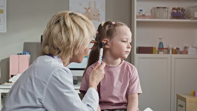 Waist up of Caucasian female pediatrician examining ear of young Caucasian child girl using otoscope in clinic. Clip perfect for pediatric healthcare marketing and medical services promotion