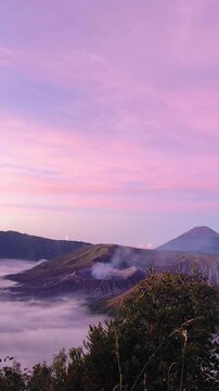 Vertical panning shot of Mount Bromo crater and Mount Semeru during a dramatic pink sunrise with a sea of clouds in Indonesia