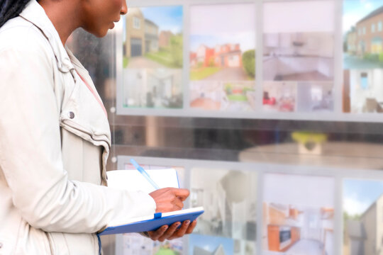 Woman writes in notebook while looking at property listings in real estate window display in a city