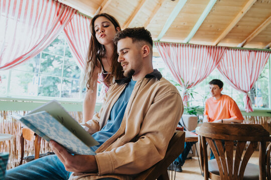 Two high school students look over a notebook while sitting in a bright cafe. A teenage girl leans in to read with a male classmate during a relaxed study moment.