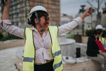 A female construction worker wearing a high-visibility vest and hard hat raises her arms in...