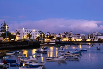 Boats float in the lagoon of Arrecife, Lanzarote as day turns to night. Buildings line the waterfront, reflecting city lights on the water. © Kotangens