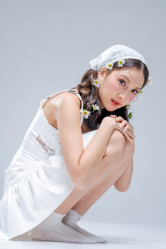 Elegant young Asian woman crouching in white satin dress with daisy flowers in hair, fashion beauty portrait hugging knees on grey background, spring bridal editorial studio shot, copy space