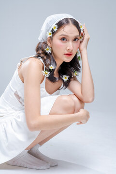 Elegant young Asian woman crouching in white satin dress with daisy flowers in hair, fashion beauty portrait hugging knees on grey background, spring bridal editorial studio shot, copy space