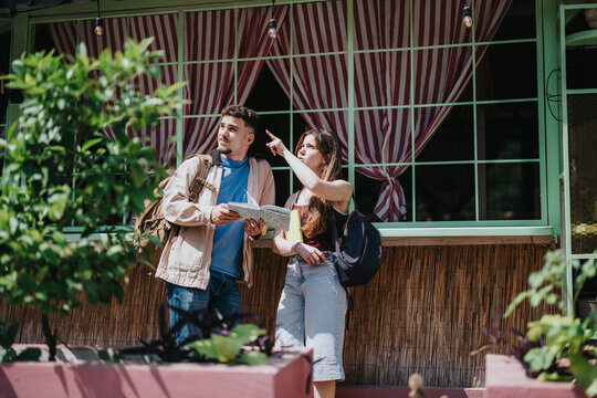 Two high school students with backpacks study a map while one points ahead. They are exploring a town and planning their route together.