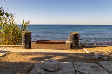 Naklejka premium Wooden bench on stone base near sea water shore view. Waterfront promenade path with black barrel planter pot in morning sun light, summer travel, old town district, Side, Turkey.