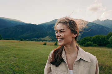 Obraz premium Smiling woman in casual shirt outdoors, bright mountains in the distance, open field under daytime sun, lifestyle portrait conveying happiness, freedom, and connection with nature.