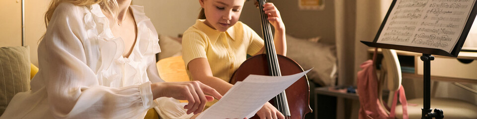Caucasian girl studying music and playing cello with female teacher, practicing reading sheet music during lesson. Ideal for music education, child learning, arts program marketing © DragonImages