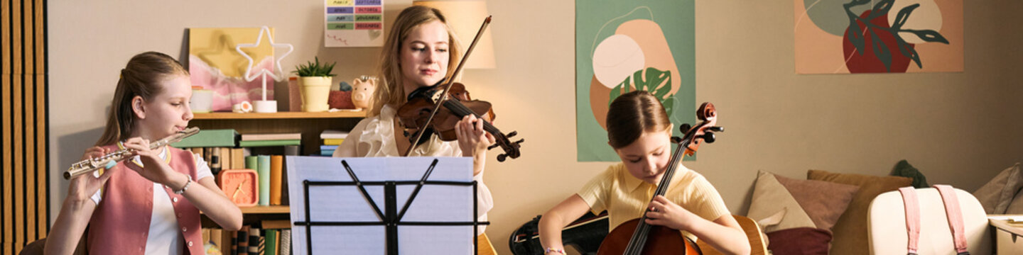 Wide shot of school age girls studying music and playing flute violin and cello at home, practicing ensemble skills and arts education. Ideal for music lessons, tutoring, youth programs