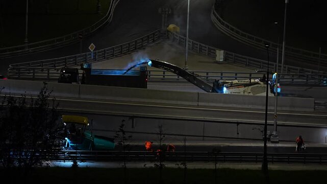Nighttime scene of road construction on an elevated overpass with illuminated heavy machinery, trucks, and workers wearing safety vests. Urban infrastructure and maintenance work

