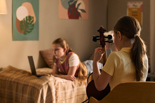 Medium shot of schoolgirl practicing cello while another kid studying music on laptop at home, supporting remote music education and online lesson marketing for learning platforms