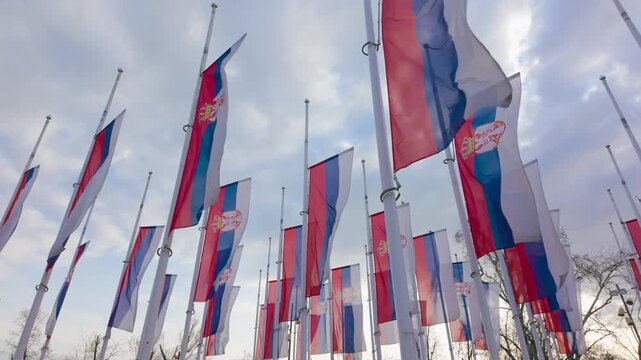 Flags flutter along tall flagpoles in an open outdoor space under a bright sky, a multicolored collection captured in wind