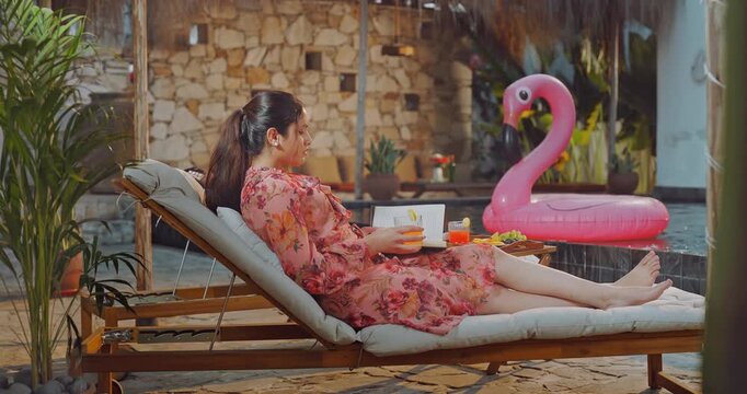 Indian young adult relax woman sitting on sun lounger chair read new novel book enjoy picnic trip day happy girl hold glass drink casual juice do self care spend time have fun joy pool edge place