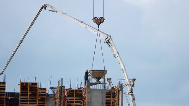 Construction site activity featuring a builder managing a concrete bucket lifted by a pump truck, pouring wet cement into a monolithic formwork for a new high rise building foundation