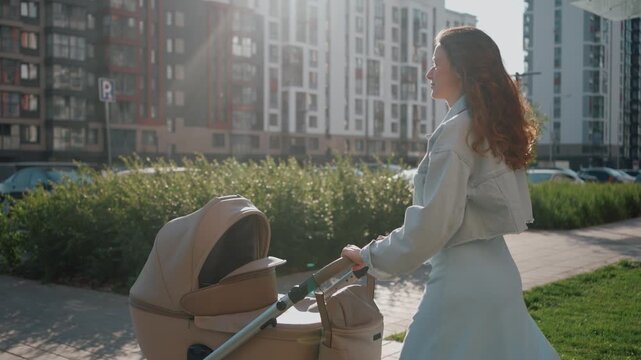 Side view tracking shot of a young redhead mother walking with a baby stroller on a sunny day. A woman pushing a pram along a sidewalk in a modern residential district with apartment buildings