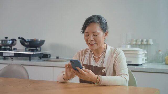 Smiling mature woman in apron watching recipes at smartphone, old housewife sitting at kitchen watch video tutorial of famous chef on phone about cooking dinner read culinary recipe at online cookbook