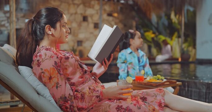Indian young adult relax woman sitting on sun lounger chair read new novel book enjoy trip day female hold glass drink juice do self care girl child eat fruits spend time have fun joy pool edge place
