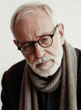 Thoughtful old man with glasses, scarf looks down with pensive gaze. His face shows wrinkles, grey beard. Calm senior man expresses deep emotion against light background. He seems to reflect on life.