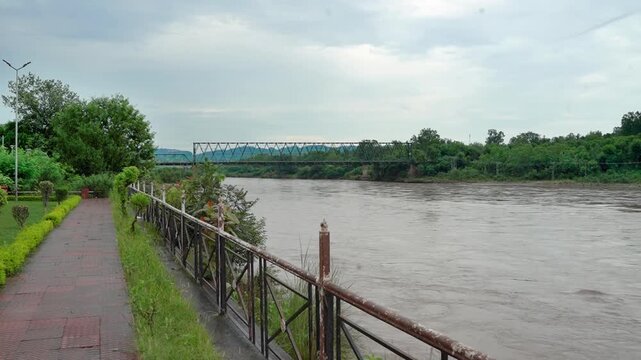 Chenab River flowing through Jammu, Jammu and Kashmir, India. The footage captures a paved riverside walking path with metal railings, a classic iron truss bridge spanning across the river,