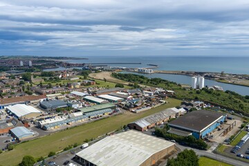 Obraz premium Aerial view across the Port of Sunderland showing harbour entrances industrial facilities warehouses riverside infrastructure and the North Sea coastline beneath dramatic cloud formations summer day