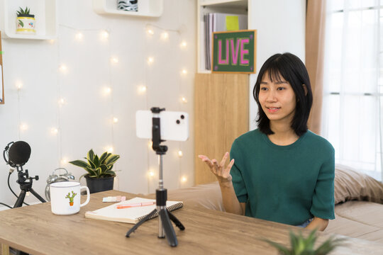 Young Creator woman live stream using a smartphone mounted on a tripod in home studio, gestures naturally while speaking with a notebook