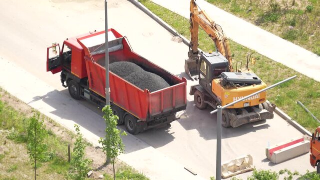 High-angle view of a red dump truck unloading black asphalt into a pile, while a yellow wheel excavator works nearby. Road construction, heavy machinery and civil engineering


