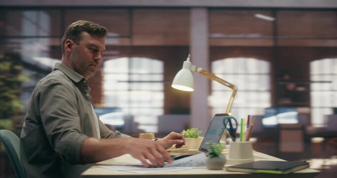 360 Degree. Caucasian Man Using Laptop Computer While Working in Brick Loft Office, Comparing Printed Spreadsheets Against Digital Analytics on Screen. Orbital Cinematic Arc Tracking Shot.